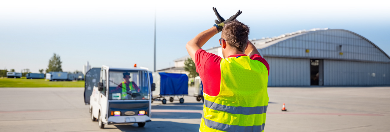 Ein Frachtabfertiger weist ein Vorfeldfahrzeug per Handzeichen sicher ein und koordiniert den Verkehr am Flughafen.