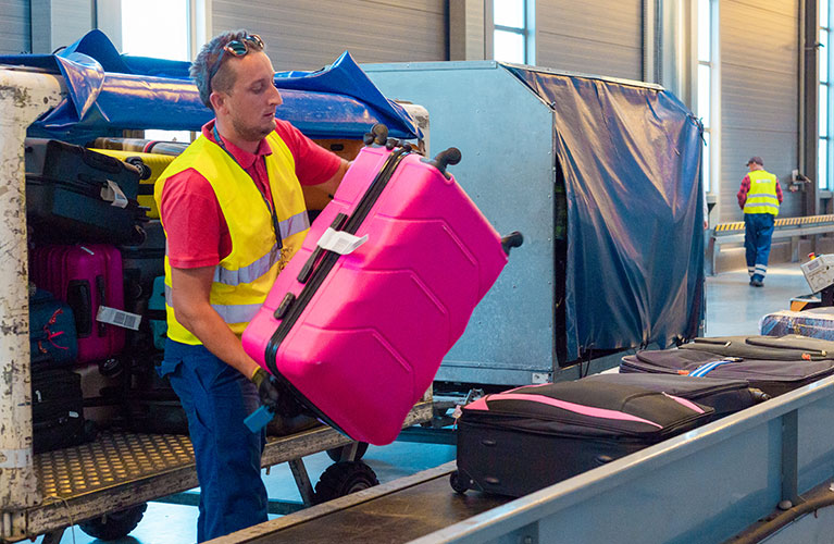 epäckabfertigung am Förderband Flughafenmitarbeiter in Warnweste hebt einen pinkfarbenen Koffer vom Gepäckwagen auf das Förderband.