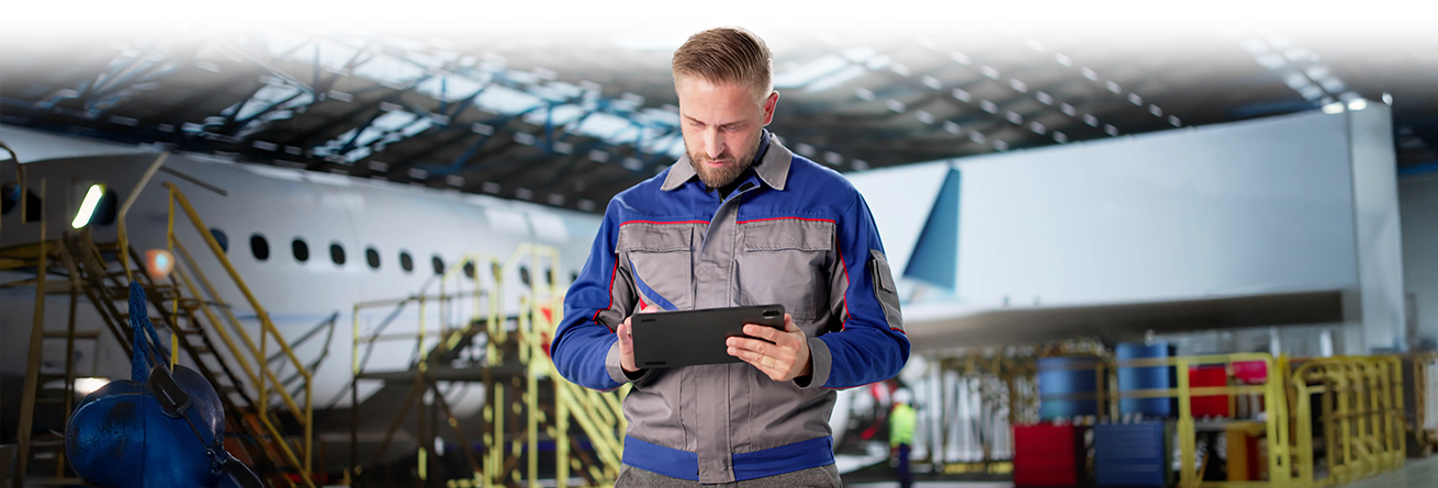 Ein Techniker in Arbeitskleidung steht in einer Flugzeughalle vor einem Flugzeug und liest oder dokumentiert Informationen auf einem Tablet.