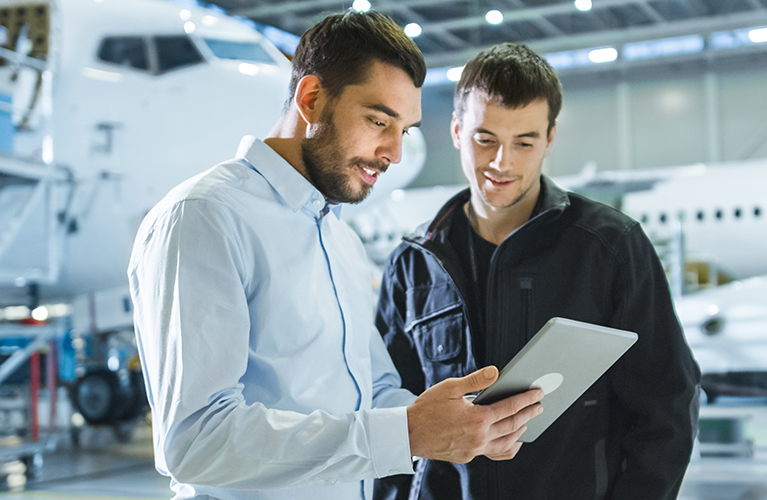 Zwei Mitarbeiter stehen in einer Flugzeughalle und betrachten gemeinsam ein Tablet. Im Hintergrund ist ein Flugzeug zu sehen, die Szene zeigt eine technische Abstimmung.