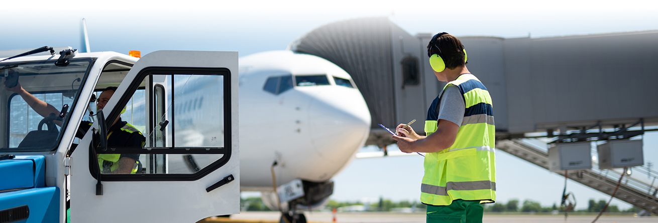 Ein Mitarbeiter mit Warnweste und Geh&ouml;rschutz steht auf dem Flughafenvorfeld vor einem Flugzeug und h&auml;lt ein Klemmbrett. Er dokumentiert oder &uuml;berpr&uuml;ft Arbeitsdaten am Flugzeug.