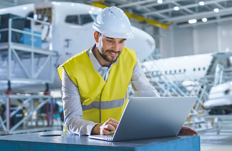 Ein Mitarbeiter mit Schutzhelm und Warnweste arbeitet in einer Flugzeughalle an einem Laptop. Im Hintergrund steht ein Flugzeug in einer Wartungsumgebung.