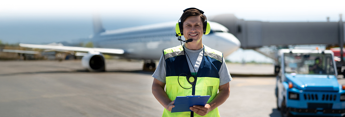 Ein Mitarbeiter mit Warnweste und Headset steht auf dem Flughafenvorfeld vor einem Flugzeug und h&auml;lt ein Klemmbrett. Im Hintergrund stehen Servicefahrzeuge.