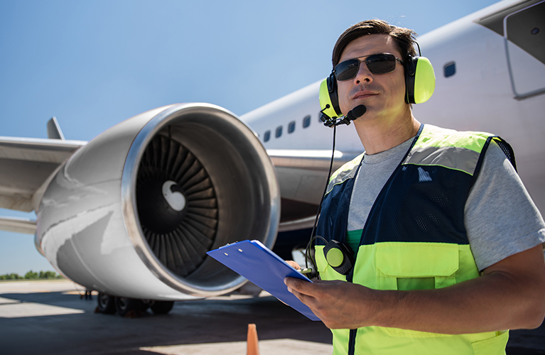 Ein Mitarbeiter mit Warnweste, Geh&ouml;rschutz und Klemmbrett steht vor dem Triebwerk eines Flugzeugs auf dem Vorfeld und &uuml;berpr&uuml;ft Daten.