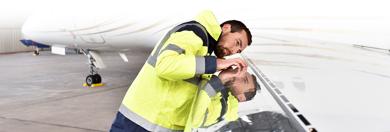 Ein Techniker in Schutzkleidung beugt sich &uuml;ber die Oberfl&auml;che eines Flugzeugs und kontrolliert diese aus n&auml;chster N&auml;he. Die Szene zeigt eine technische Inspektion.