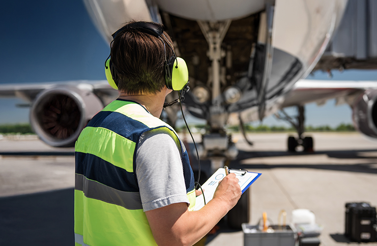 Ein Mitarbeiter mit Warnweste und Geh&ouml;rschutz steht vor einem Flugzeug und schreibt auf einem Klemmbrett. Das Flugzeugfahrwerk ist im Hintergrund zu sehen.