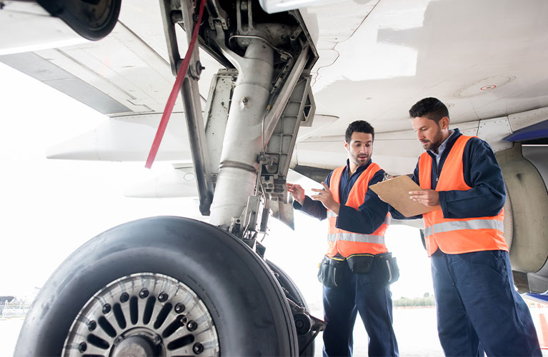 Zwei Flugger&auml;tmechaniker kontrollieren das Fahrwerk eines Flugzeugs, dokumentieren Pr&uuml;fergebnisse und stellen die technische Sicherheit vor dem Einsatz sicher.