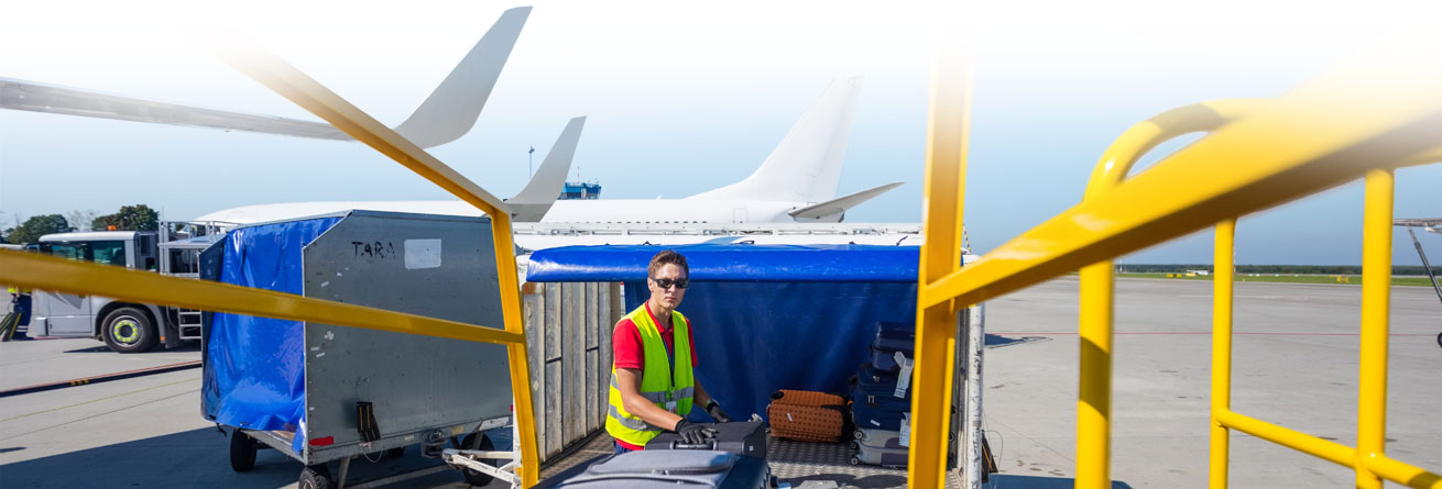 Gepäckverladung am Rollfeld Flughafenmitarbeiter in Warnweste lädt Koffer von einem Gepäckwagen auf das Förderband, im Hintergrund Flugzeuge.