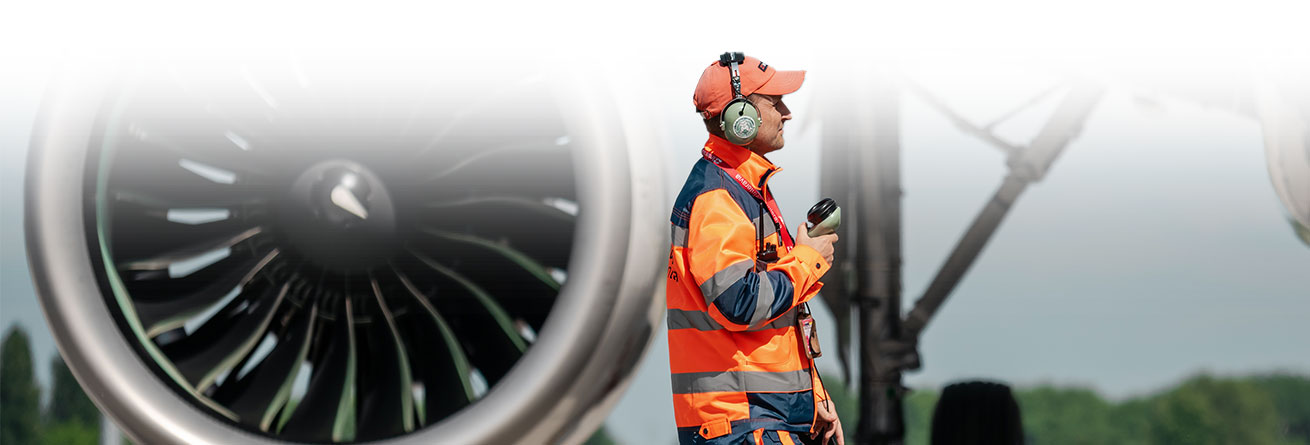 Ramp Agent in orangener Warnkleidung mit Funkgerät steht vor einem Flugzeugtriebwerk.