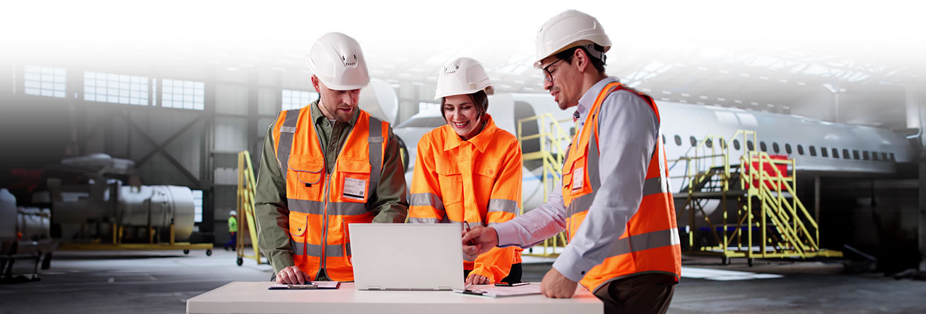 Teamarbeit in der Flugzeugwartungshalle Drei Mitarbeiter in orangenen Warnwesten und Schutzhelmen besprechen Pläne an einem Laptop in einer Flugzeughalle.