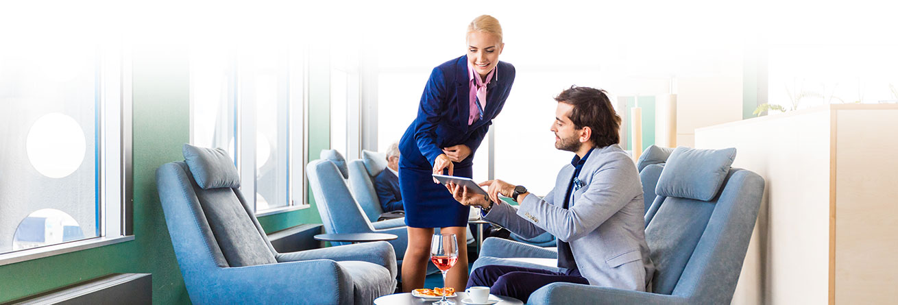 Flughafenmitarbeiterin in Uniform berät einen Passagier in der Lounge, während Getränke serviert werden.