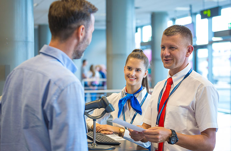 Zwei Mitarbeitende in Uniform stehen an einem Serviceschalter im Flughafen und sprechen mit einem Reisenden. Sie halten Unterlagen bereit und unterst&uuml;tzen ihn beim Check-in oder einem Servicethema. Die Umgebung ist hell und modern gestaltet.