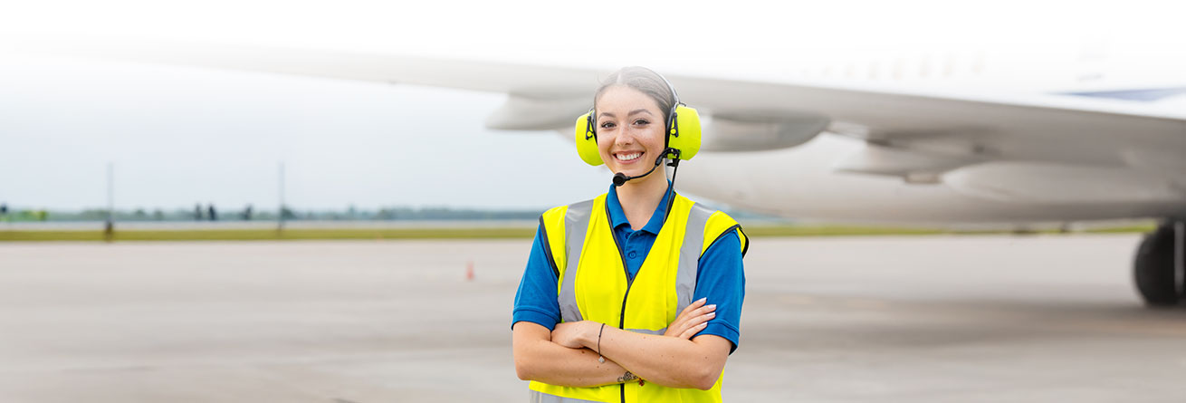 Eine Person in Warnweste, Geh&ouml;rschutz und Headset steht l&auml;chelnd vor einem Flugzeug auf dem Rollfeld. Die Person hat die Arme verschr&auml;nkt und tr&auml;gt Arbeitskleidung f&uuml;r den Einsatz im Au&szlig;enbereich.