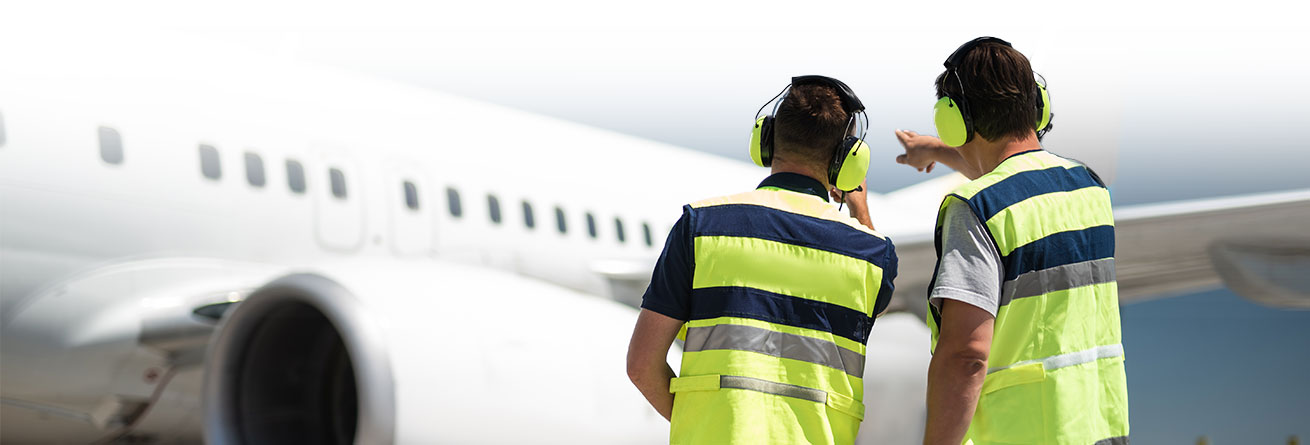 Zwei Flughafenmitarbeiter mit Gehörschutz und Warnwesten stehen vor einem Flugzeug und weisen den Weg ein.
