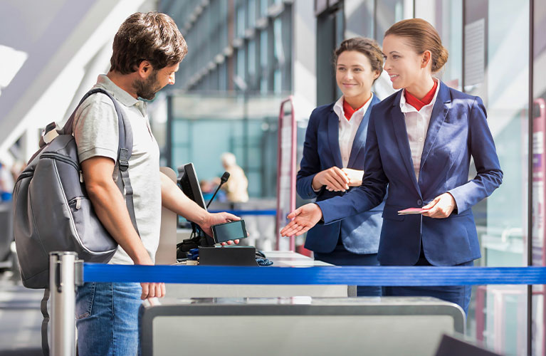 Zwei Mitarbeitende in Uniform stehen an einem Check-in- oder Kontrollschalter und unterst&uuml;tzen einen Reisenden, der sein Smartphone oder ein Ticket vorzeigt. Die Szene spielt in einer modernen, hellen Flughafenhalle.