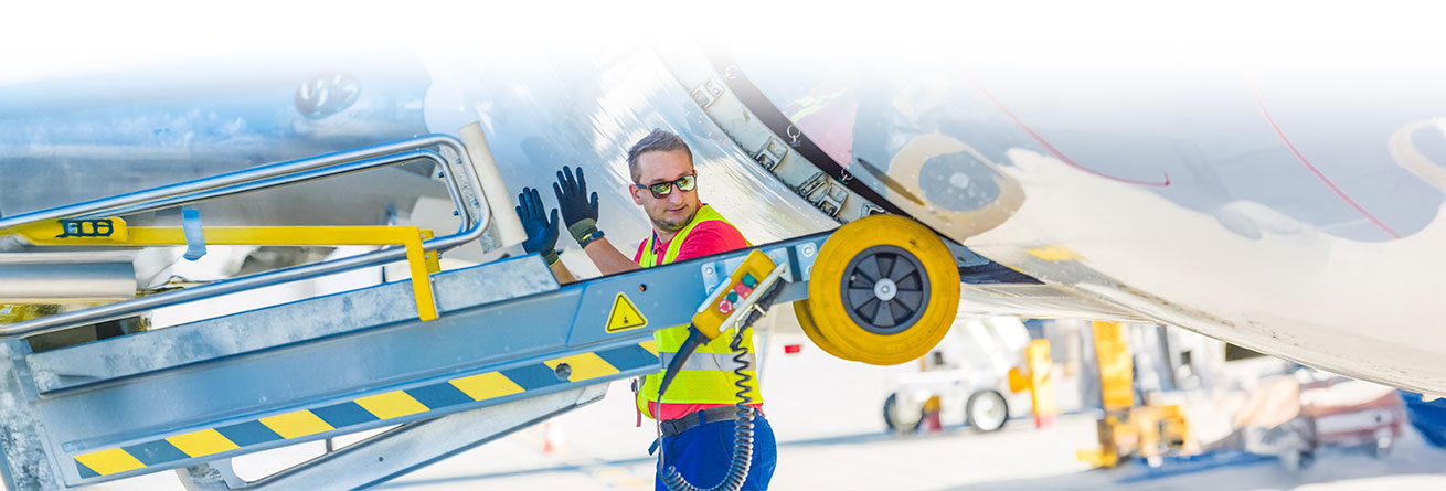 Eine Person in Warnweste und Schutzausr&uuml;stung arbeitet an einem Flugzeug. Sie steht an einer mobilen Rampe oder einem Bodenfahrzeug und ber&uuml;hrt die Unterseite des Flugzeugs, w&auml;hrend sie den Arbeitsbereich &uuml;berwacht.