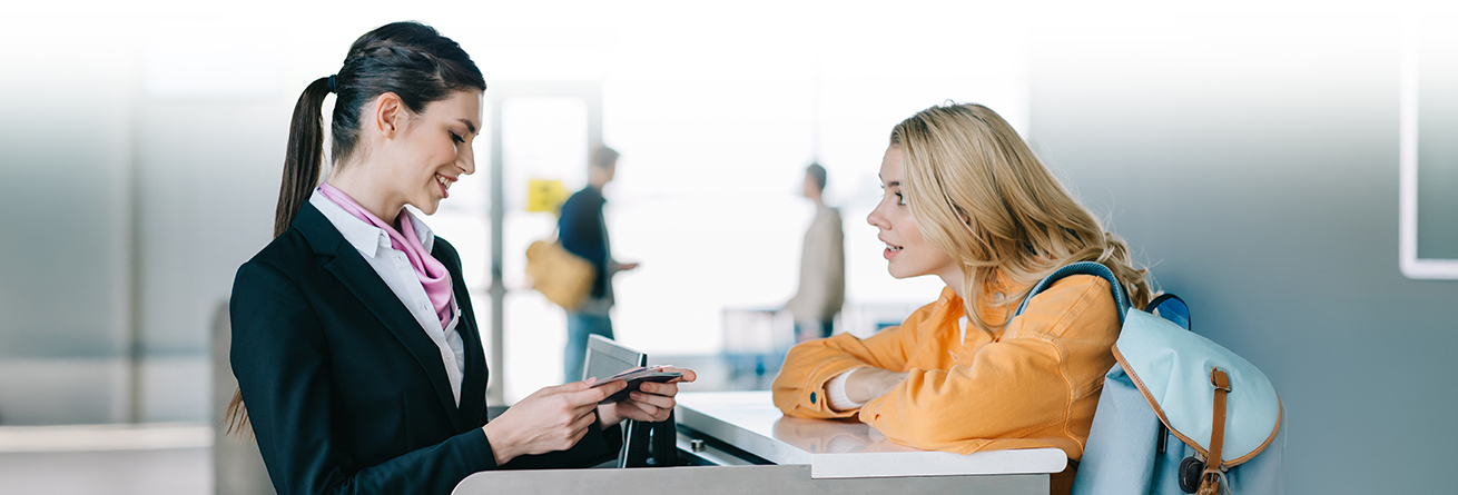 Eine Mitarbeiterin am Flughafenschalter pr&uuml;ft die Reisedokumente einer Reisenden mit Rucksack. Beide stehen sich am Counter gegen&uuml;ber, im Hintergrund sind weitere Passagiere unscharf zu erkennen.