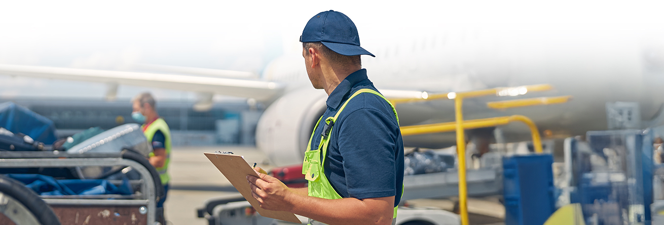 Ein Mitarbeiter mit Warnweste und Klemmbrett steht auf dem Flughafenvorfeld neben Gep&auml;ckwagen. Im Hintergrund ist ein Flugzeug zu sehen, w&auml;hrend weiteres Bodenpersonal arbeitet.