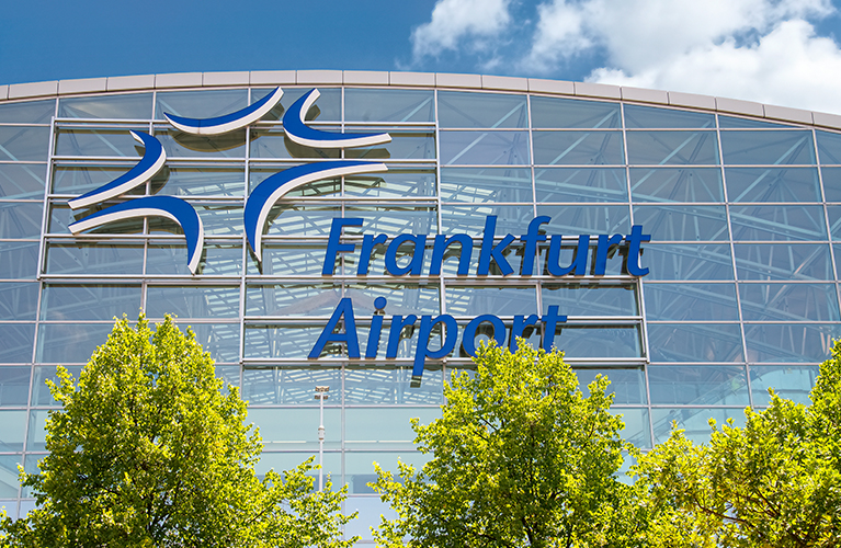 Die Glasfassade eines Terminalgeb&auml;udes mit dem Schriftzug &bdquo;Frankfurt Airport&ldquo;. Vor dem Geb&auml;ude stehen gr&uuml;ne B&auml;ume, dar&uuml;ber ist ein blauer Himmel zu sehen.