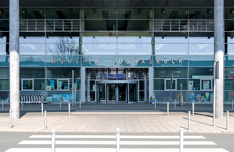 Die Frontansicht des Bahnhofs M&uuml;nster (Westfalen) mit gro&szlig;em Glas-Eingang und automatischen T&uuml;ren. Vor dem Geb&auml;ude befinden sich ein Vorplatz mit Fahrbahnmarkierungen sowie Fahrradst&auml;nder.